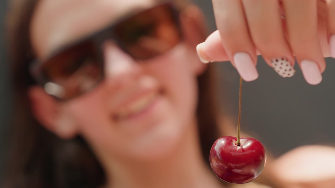Donna sorridente tiene una singola ciliegia rossa vicino alla fotocamera, mettendo in mostra le sue unghie curate e gli occhiali da sole. L'attenzione rimane sulla mano che tiene il frutto lucido su uno sfondo sfocato.