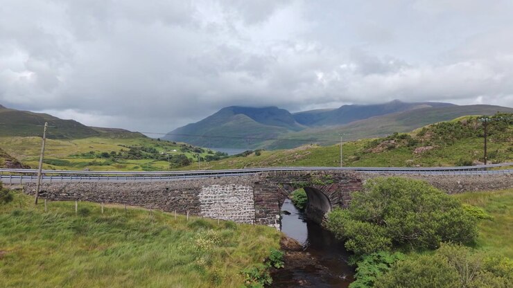 Un autentico ponte ad arco in pietra attraversa un piccolo fiume nelle lussureggianti colline verdi della Contea di Mayo, Irlanda. Un'auto attraversa la strada moderna sovrastante