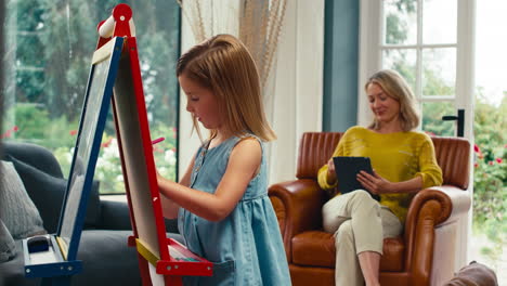 family with daughter drawing picture on easel as mother with digital tablet watches from sofa