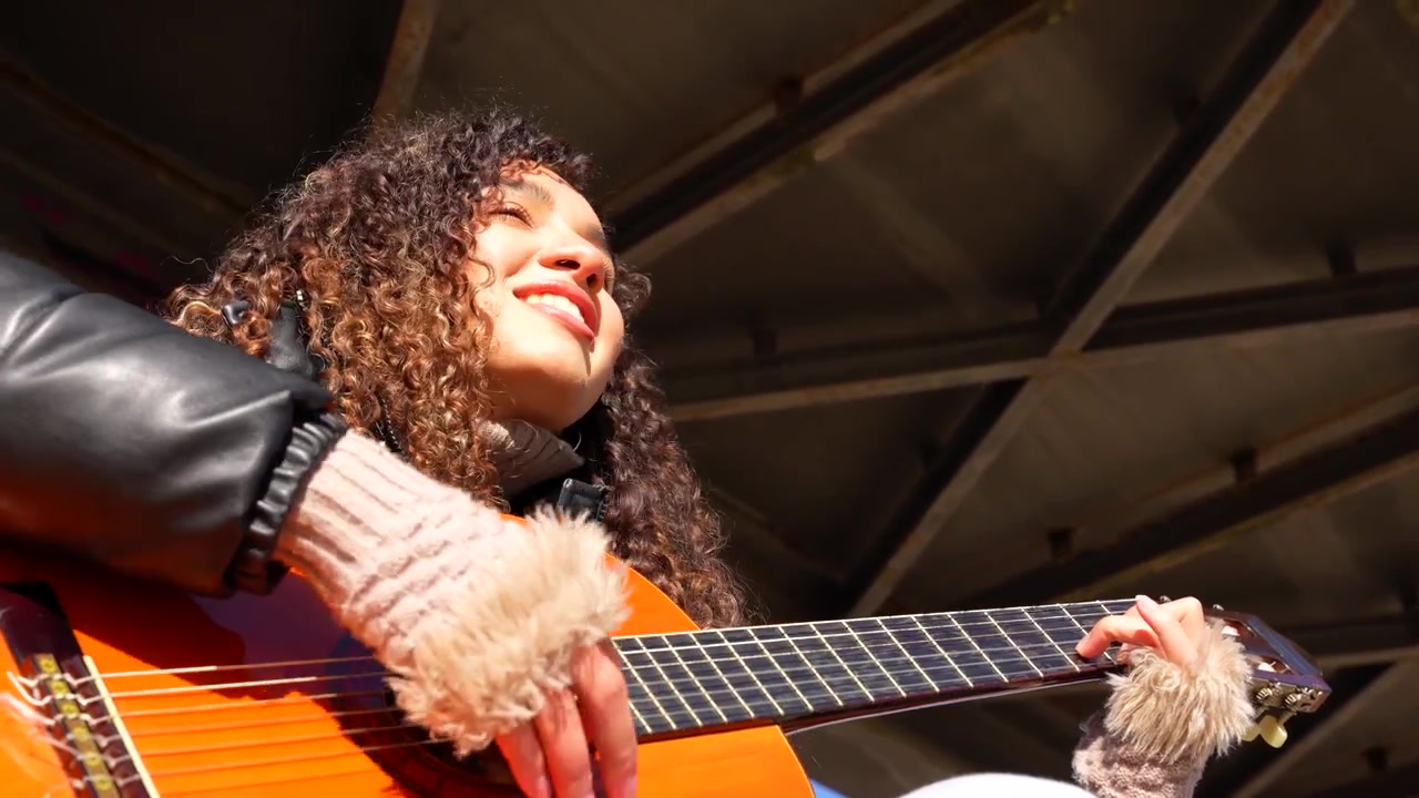 Ung krølhåret kvindelig musiker spiller akustisk guitar i bymiljø under en bro