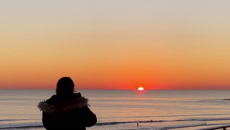 mujer joven tomando fotografías con su teléfono inteligente en la puesta de sol en la playa y rompiendo olas