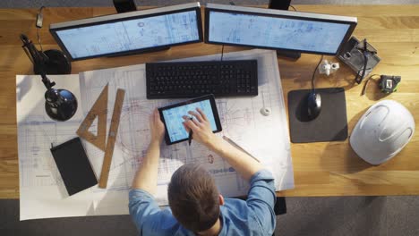 top view of architectural engineer draws on his blueprints, compares with tablet computer with green screen, using desktop computer also. his desk is full of useful objects and evening sun.