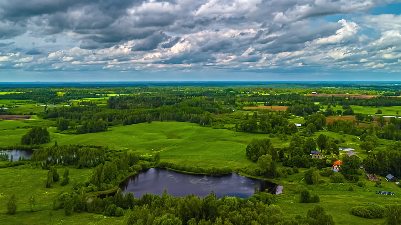 Stunning aerial time-lapse showcasing picturesque Latvian farmlands with serene lakes, scattered forests, lush green meadows, and charming small cottage houses under a sky of moving clouds