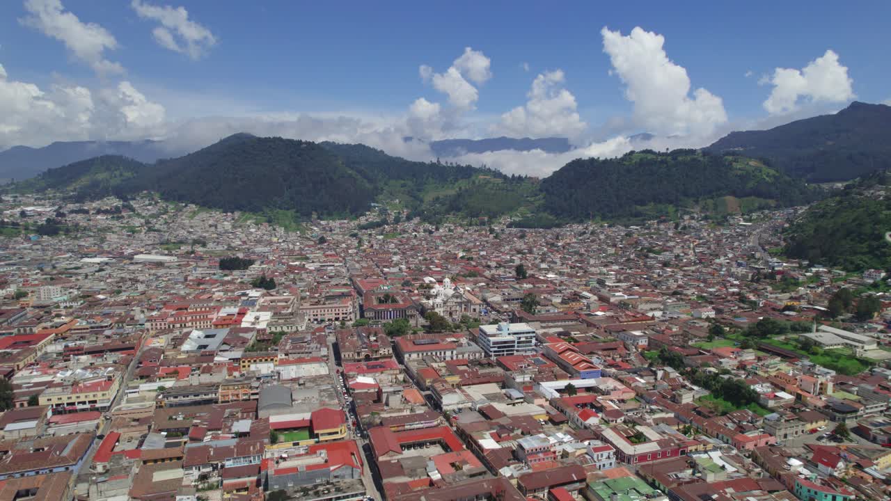 Drone aerial footage of urban central american city Quetzaltenango, Xela, Guatemala showing colorful red rooftops and cityscape surrounded by mountains. Zone 1 and Zone 4. Bright partly cloudy day.