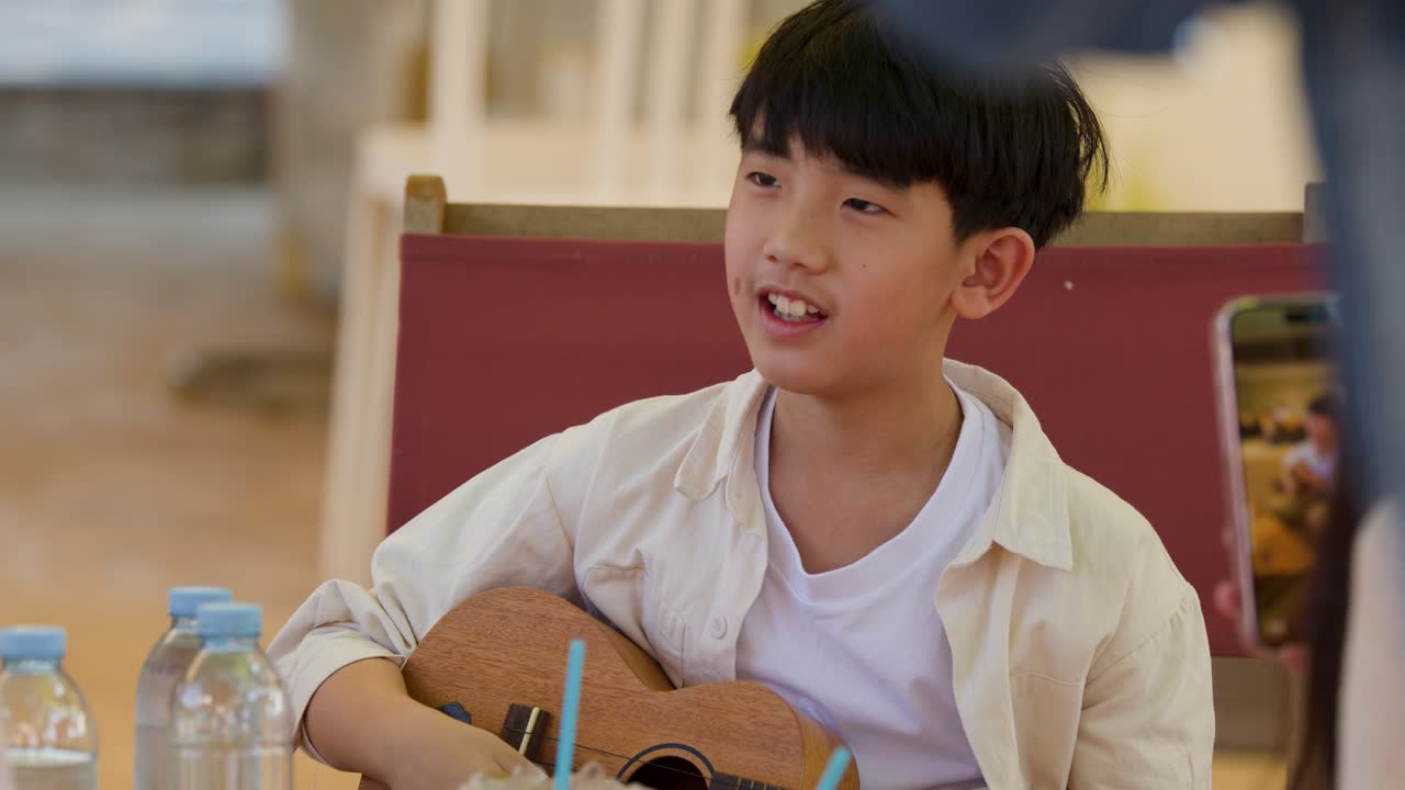 Un giovane ragazzo thailandese siede all'aperto, concentrato a suonare un ukulele durante una lezione di musica.