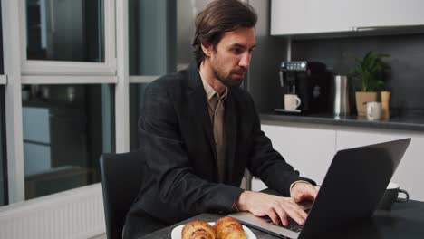 a serious and confident brunette man in a black jacket and shorts works at a gray laptop while sitting at the dining table in the kitchen working remotely in a modern apartment