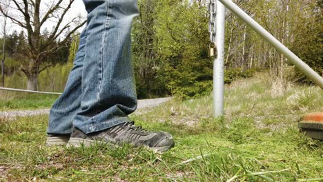 vista del tobillo de una persona que usa jeans cortando hierba verde con un devorador de malezas por una encuesta de acero metálico en el campo