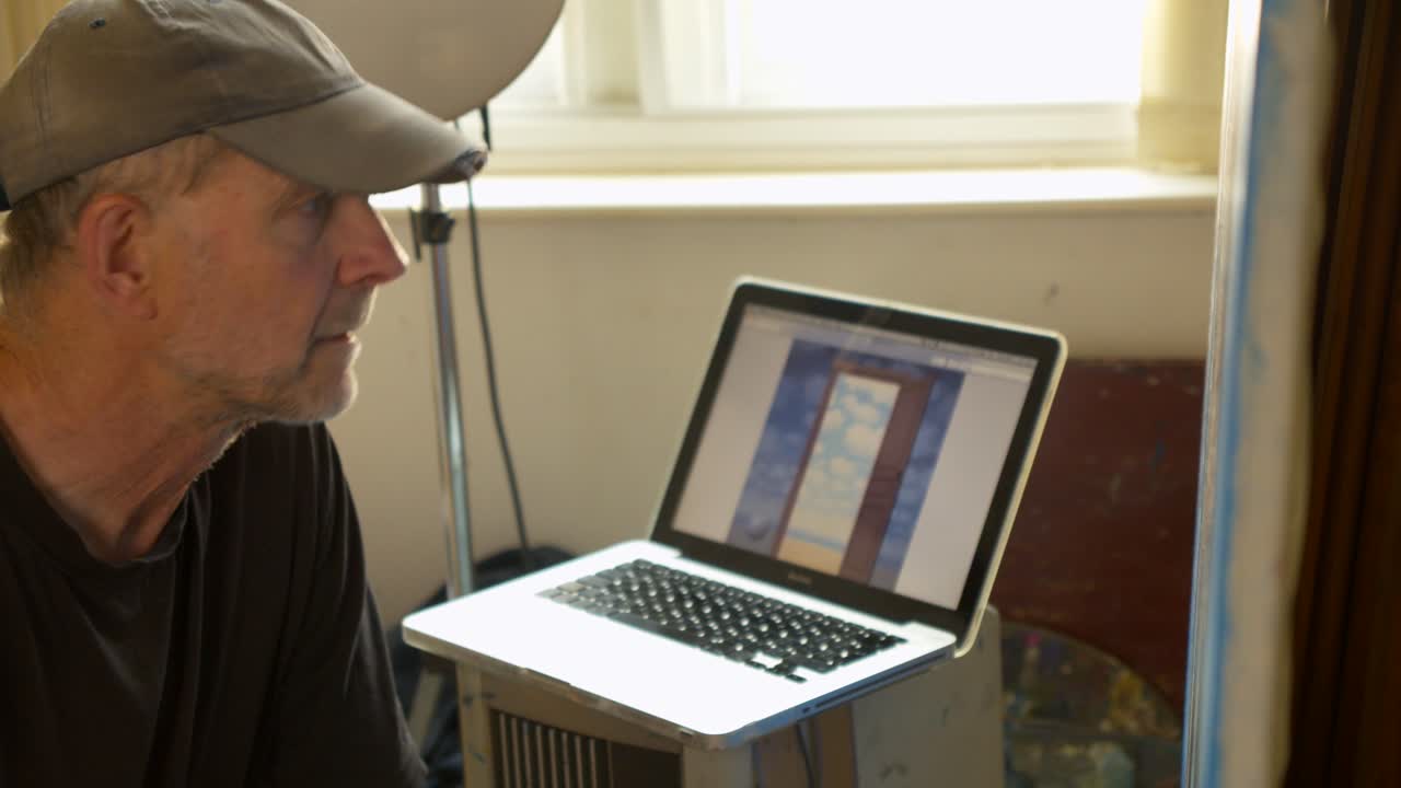 un retratista masculino, de perfil, hace referencia a una computadora portátil mientras pinta un lienzo de trabajo