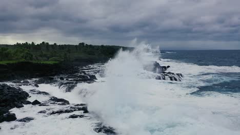 A-massive-wave-crashes-onto-rocky-shores-in-Hawaii,-with-lush-green-vegetation-and-a-dramatic-cloudy-sky-in-the-background