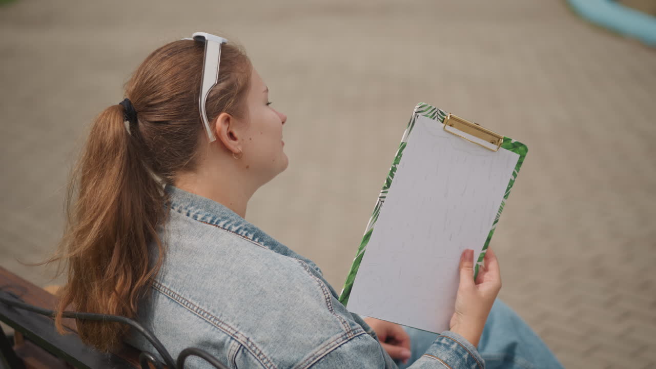 Ung kaukasisk kvinde holder et tomt clipboard, stirrer ud i det fjerne, designer planlægger layout, sidder på en bænk, denimjakke, eftertænksom positur, bysti, bløde neutrale toner