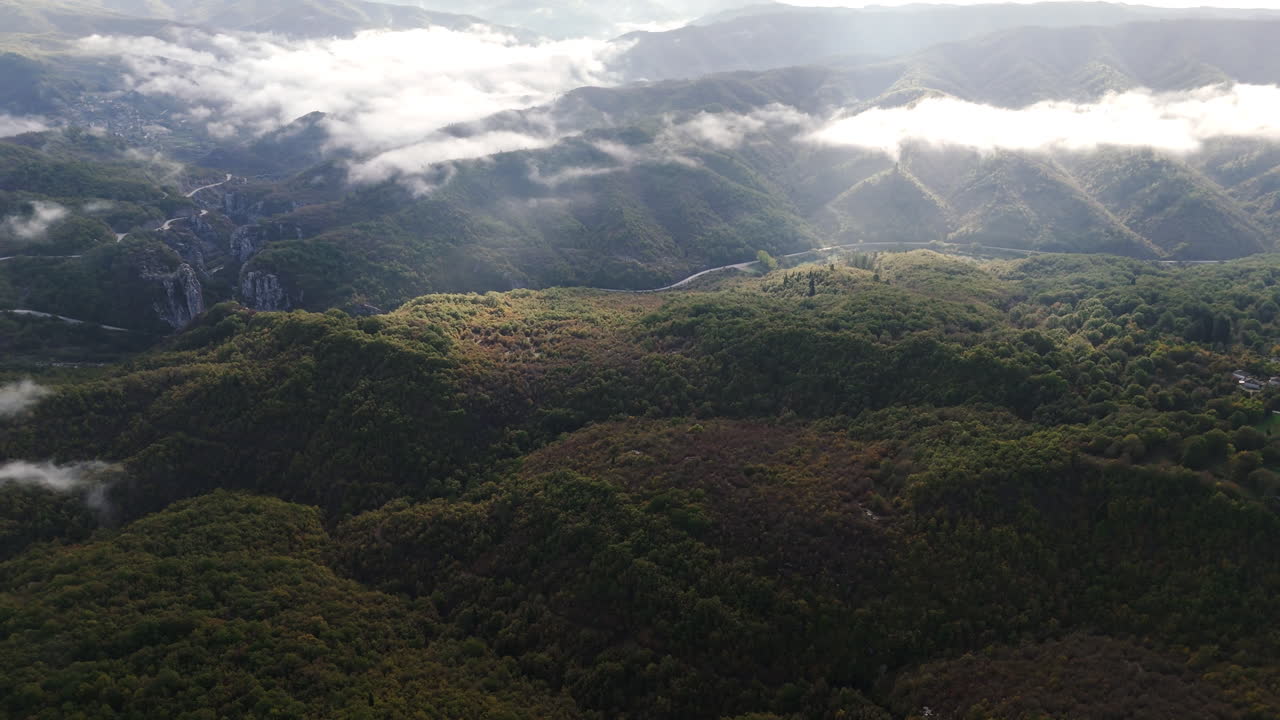 Aerial overview of central Zagori with expansive misty valleys and winding mountain roads as sunlight breaks through cloud shadows onto forest