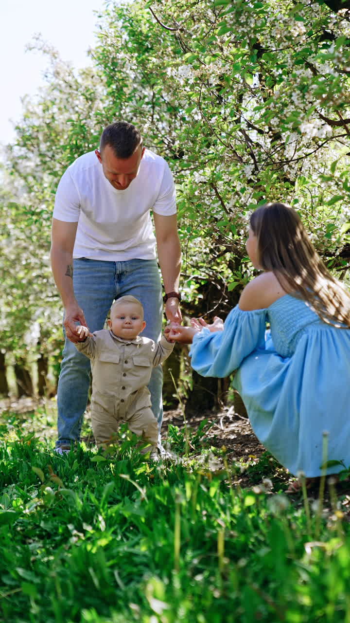 Smiling Caucasian man leading his infant baby by the hands in the garden. Mother in blue dress calls up her beloved child. Vertical video.