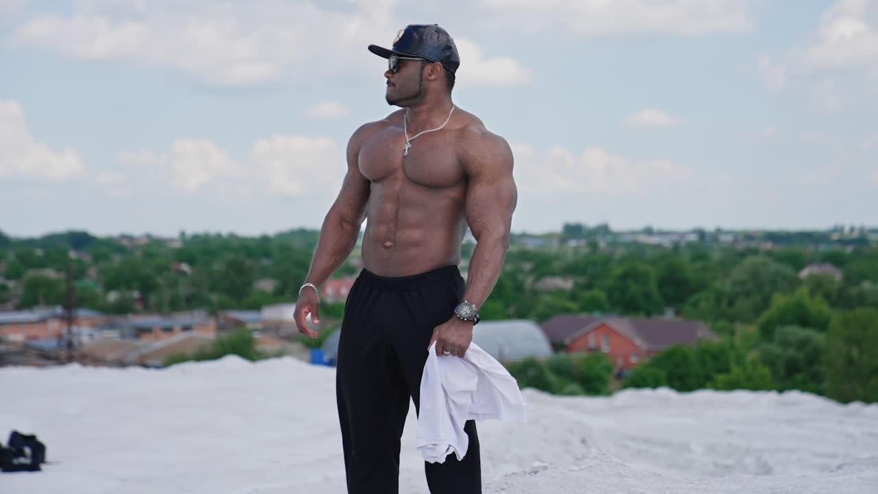 Shirtless athlete with dark skin outdoors. Muscular man standing on white hill in the countryside. Handsome sportive african american guy without shirt looks aside.
