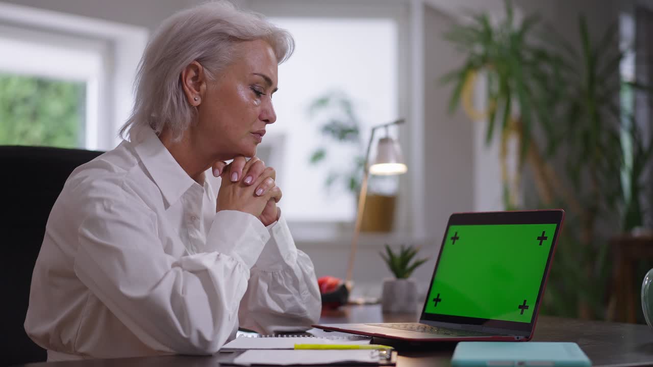 vista lateral concentrada mujer de negocios de mediana edad sentada en la mesa con chromakey computadora portátil pensando. enfocada exitosa hermosa mujer caucásica viendo noticias analizando el comercio electrónico en el interior
