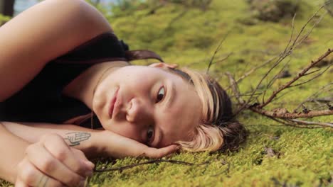 a beautiful young adult woman is lying on green and mossy forest ground while enjoying sunshine and calmness