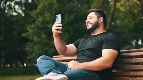 Hombre-Guapo-Sentado-En-Un-Banco-En-El-Parque-Riendo-Y-Charlando-A-Través-De-Una-Videollamada-Con-Teléfono-Celular