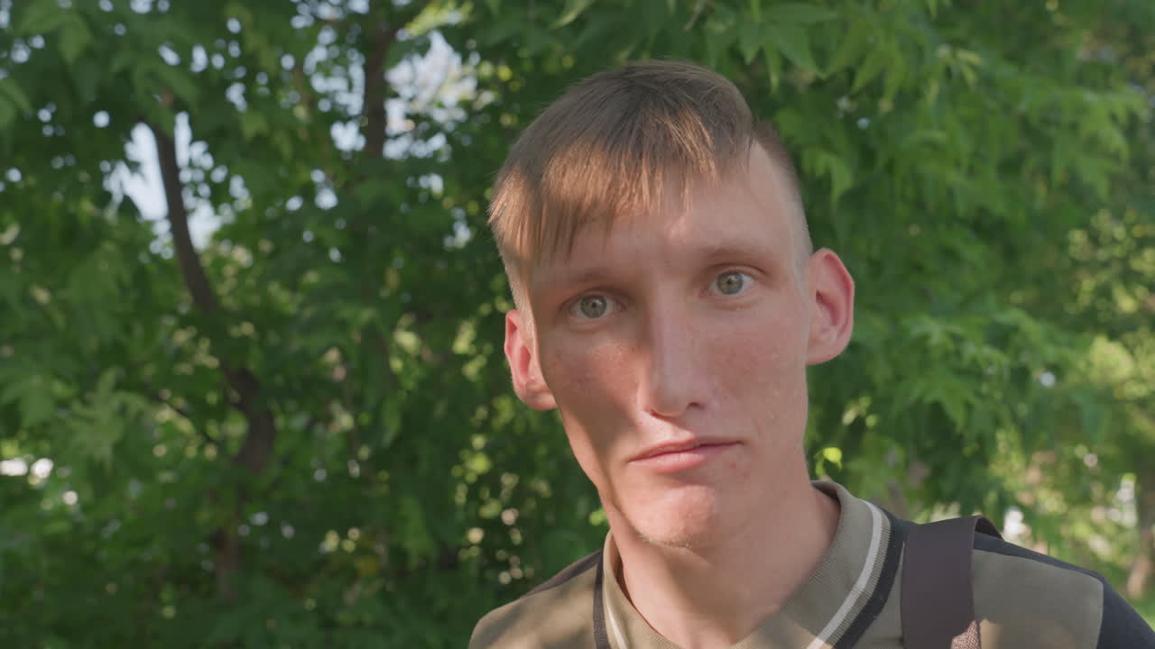 CloseUp Outdoor Portrait Under Green Trees, Young Man Stares With Tense Expression, Backpack Strap Visible, Dappled Sunlight And Leafy Background, Candid Handheld Framing, Subtle Shadow Across Face