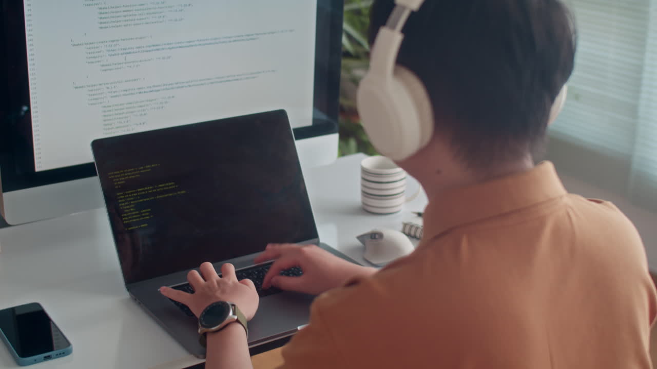 Programmer in Headphones Coding on Laptop in front of Big Monitor at Office