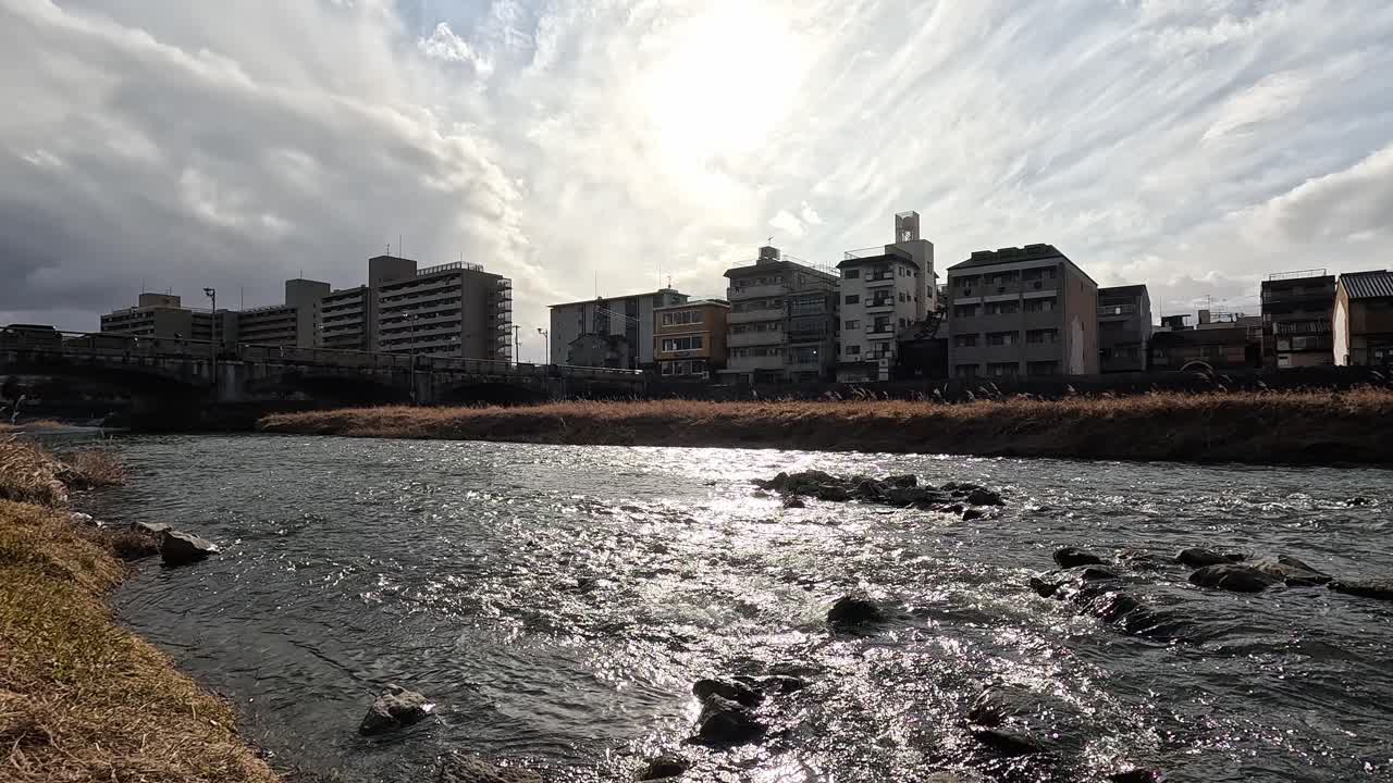 Sunlight reflects off the flowing Kamo River in Kyoto with urban buildings and dramatic clouds in the background.