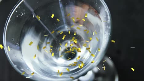 Animation-of-gold-confetti-falling-over-olives-dropping-into-clear-drink-in-glass,-on-black