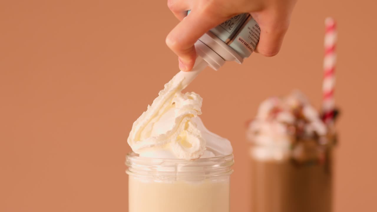 Primer plano de una mano vertiendo nata montada en un batido de vainilla, con un batido de chocolate desenfocado al fondo