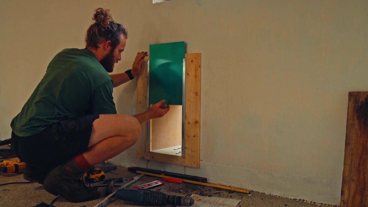 Un uomo sta forando e regolando un cancelletto per galline sopra un'apertura nel muro, con attrezzi e strumenti di misurazione sparsi intorno a lui - Primo piano.