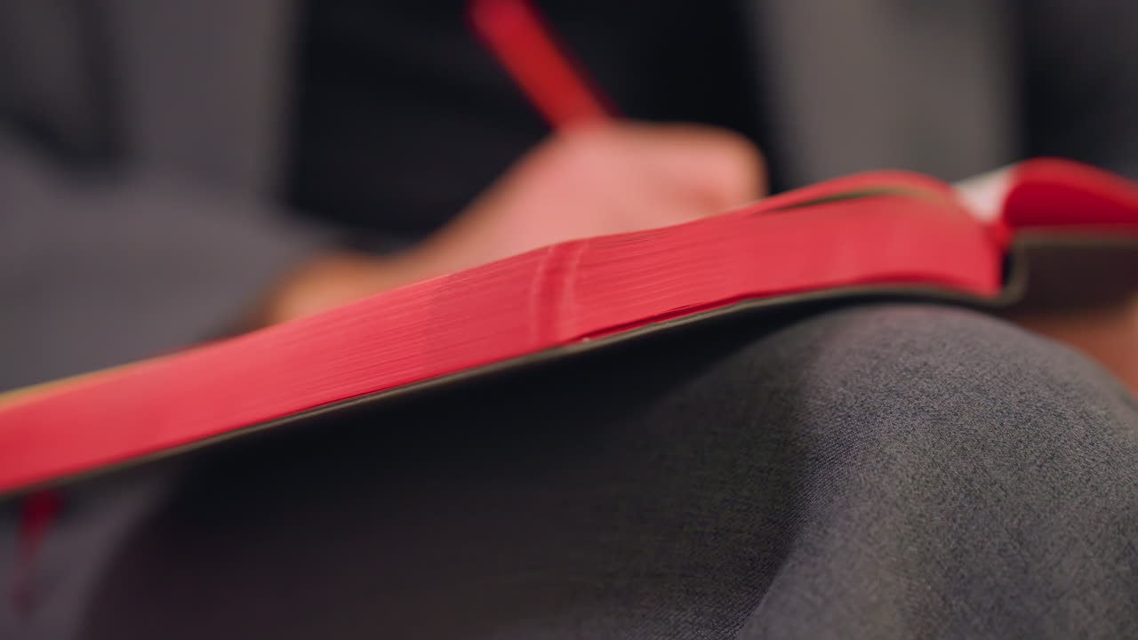 Close-up of businesswoman s hand writing in red notebook with red pen, part of body blurred, focusing on hand movement and writing in office setting, representing productivity and professional work