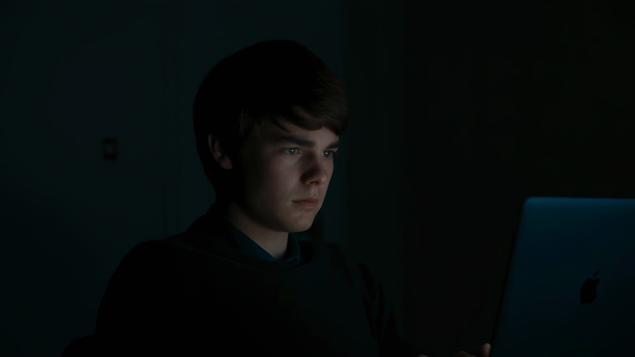 White Teen Staring At Laptop In Dark Room Lit By Blue Screen Glow, Latenight Coder Studying With Focused