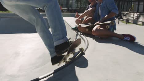 una mujer y un hombre caucásicos riendo sentados, mirando a un amigo patinando en un día soleado