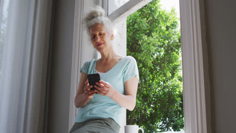 Mujer-Caucásica-Mayor-Usando-Un-Teléfono-Inteligente-Cerca-De-La-Ventana-En-Casa