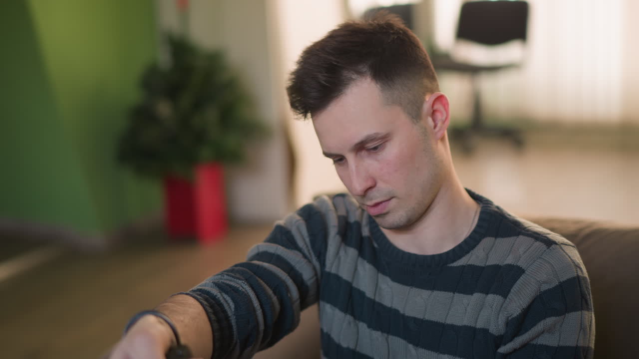Uomo concentrato che regola una telecamera o attrezzatura per le riprese mentre è seduto sul divano in salotto, preparandosi per la creazione di video o contenuti. Ambiente interno informale con illuminazione naturale.
