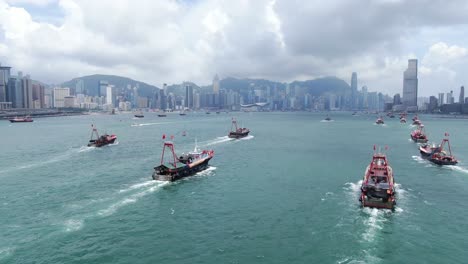 Convoy-of-local-Fishing-boats-causing-in-Hong-Kong-Victoria-bay,-with-city-skyline-in-the-horizon,-Aerial-view