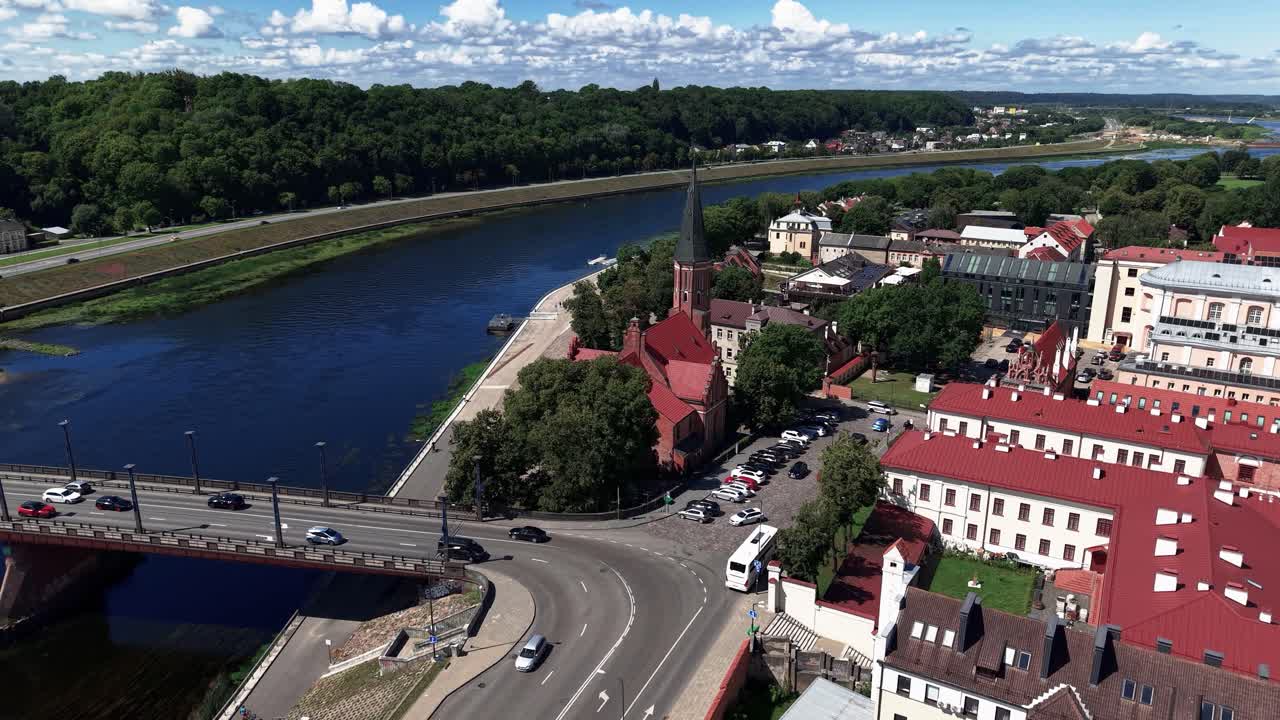 Aerial view of a scenic European city with a red-roofed old town, church spire, river, and bridge on a sunny day. Traffic flows along the road near historic buildings