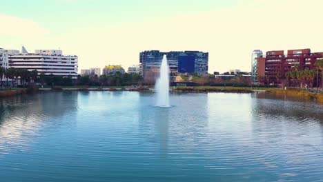 vuelo de drones sobre un lago con una fuente en medio de la ciudad, rodeado de vegetación, salones y edificios residenciales modernos
