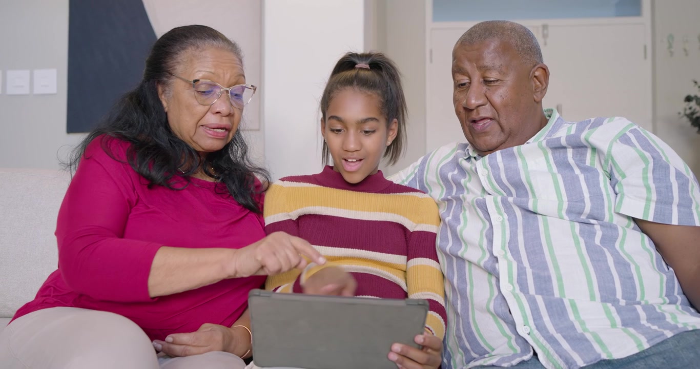 African American family leaning forward on sofa, youth girl holding tablet guiding seniors tapping