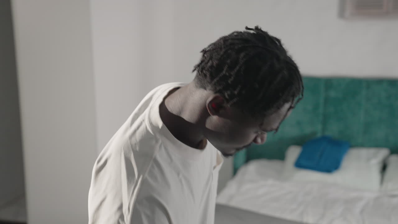 Man in white t-shirt looking down in minimalist bedroom, holding backpack, with green headboard, bed, and simple decor in background, preparing to leave, peaceful and clean home environment