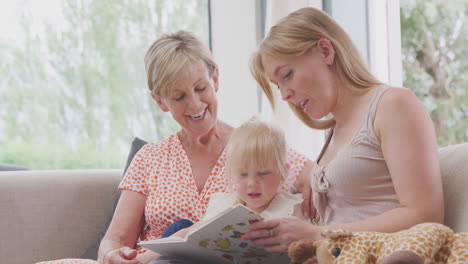 nieta sentada en el sofá con la madre y la abuela en casa leyendo un libro juntos