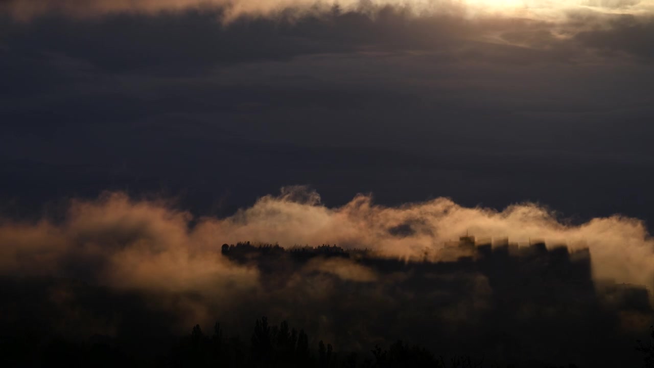Časosběr dramatických mraků a mlhy valících se přes městské panorama při východu slunce, osvětlených teplým zlatavým světlem.