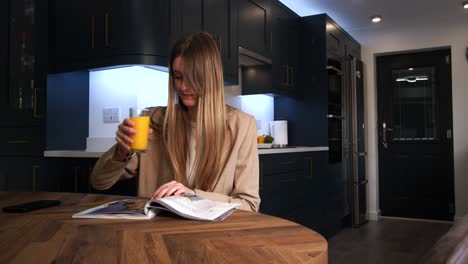 modelo femenina con una chaqueta inteligente bebiendo de un vaso de jugo de naranja leyendo una revista y mirando el teléfono en una mesa de cocina en una cocina moderna