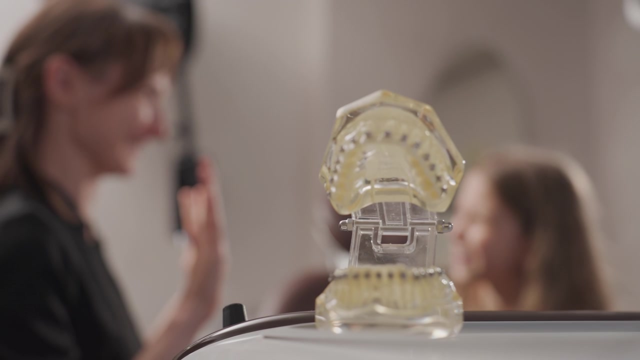 Primo piano di una donna e una bambina durante una consultazione ortodontica. L'adulto spiega il trattamento dentale tenendo in mano gli strumenti, mentre la bambina ascolta attentamente vicino all'attrezzatura specializzata. Illuminazione soffusa.