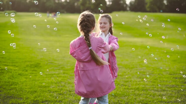 Sorelline felici con vestiti identici circondate da bolle di sapone, che si tengono per mano e volteggiano nel parco