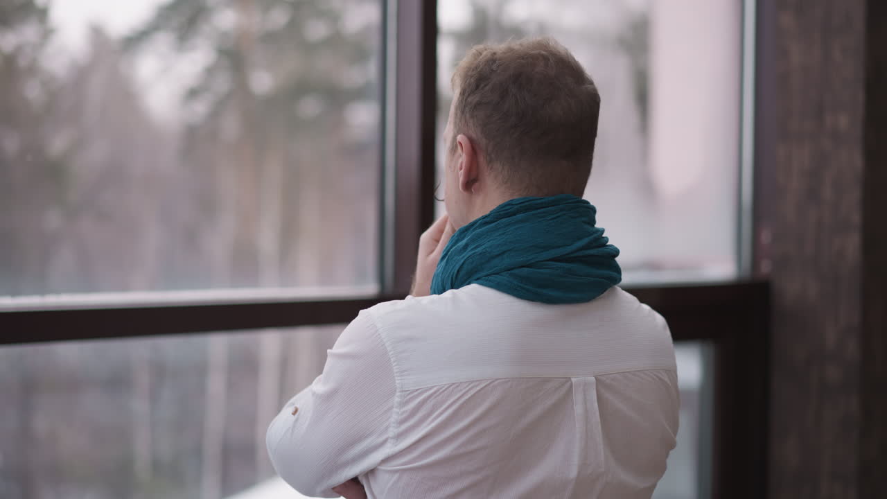 스카프를 입은 남자는 겨울의 풍경을 바라보고, 집에서 창문 근처에서 사려 깊은 고려로 문제를 해결하는 데 관심을 기울인다.