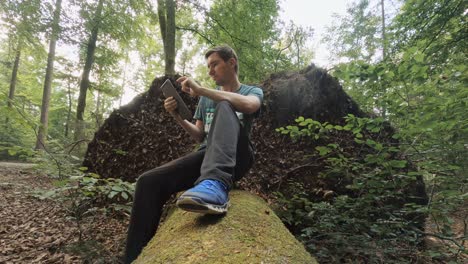 A-man-is-sitting-in-a-green-forest-at-a-fallen-tree-trunk-and-is-typing-something-at-his-tablet-computer---enjoy-working-at-the-open-fresh-air