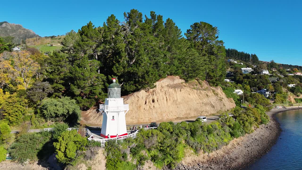Aerial footage captures Akaroa Lighthouse surrounded by lush greenery and calm waters under clear blue skies