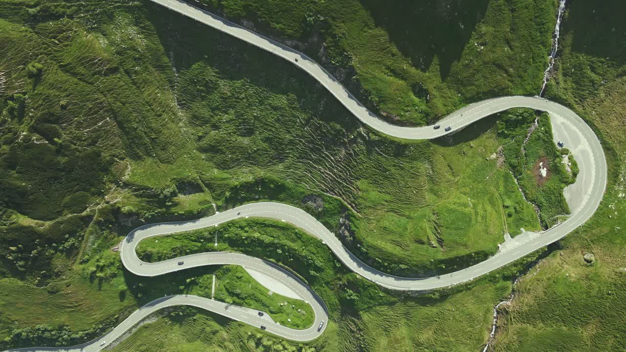 An aerial video shows a serpentine mountain road snaking through a lush, green landscape. Multiple switchbacks and vehicles are visible, emphasizing the road's dramatic curves and elevation.