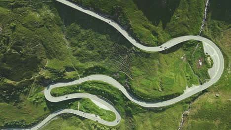 an aerial video shows a serpentine mountain road snaking through a lush, green landscape. multiple switchbacks and vehicles are visible, emphasizing the road's dramatic curves and elevation.