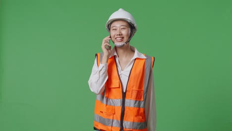 안전 헬을 입은 아시아 여성 엔지니어가 녹색 화면 배경 스튜디오에서 는 동안 스마트폰으로 이야기합니다.