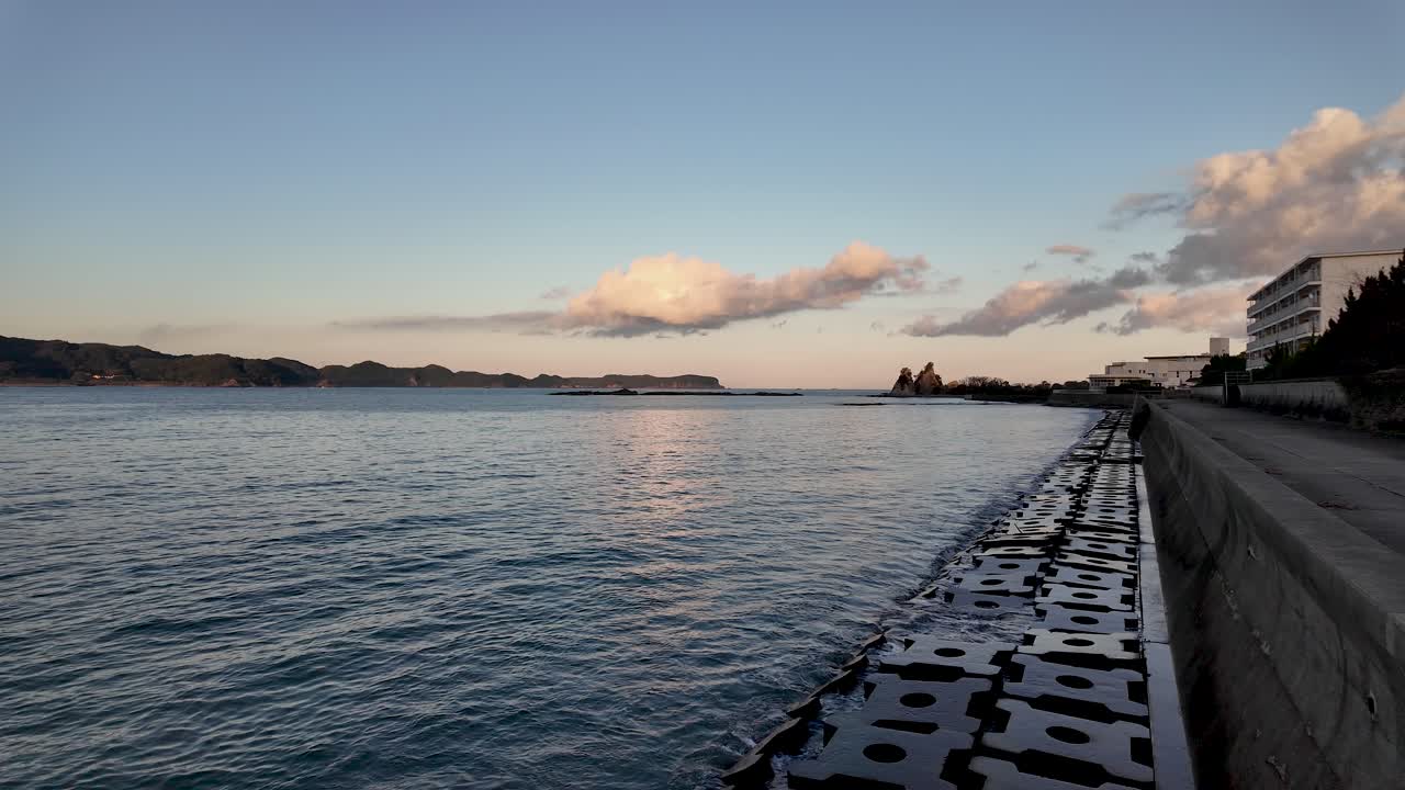 Tranquil scene of Nachi Bay during golden hour, featuring a breakwater wall and serene ocean waters under a sky with soft clouds. slow motion