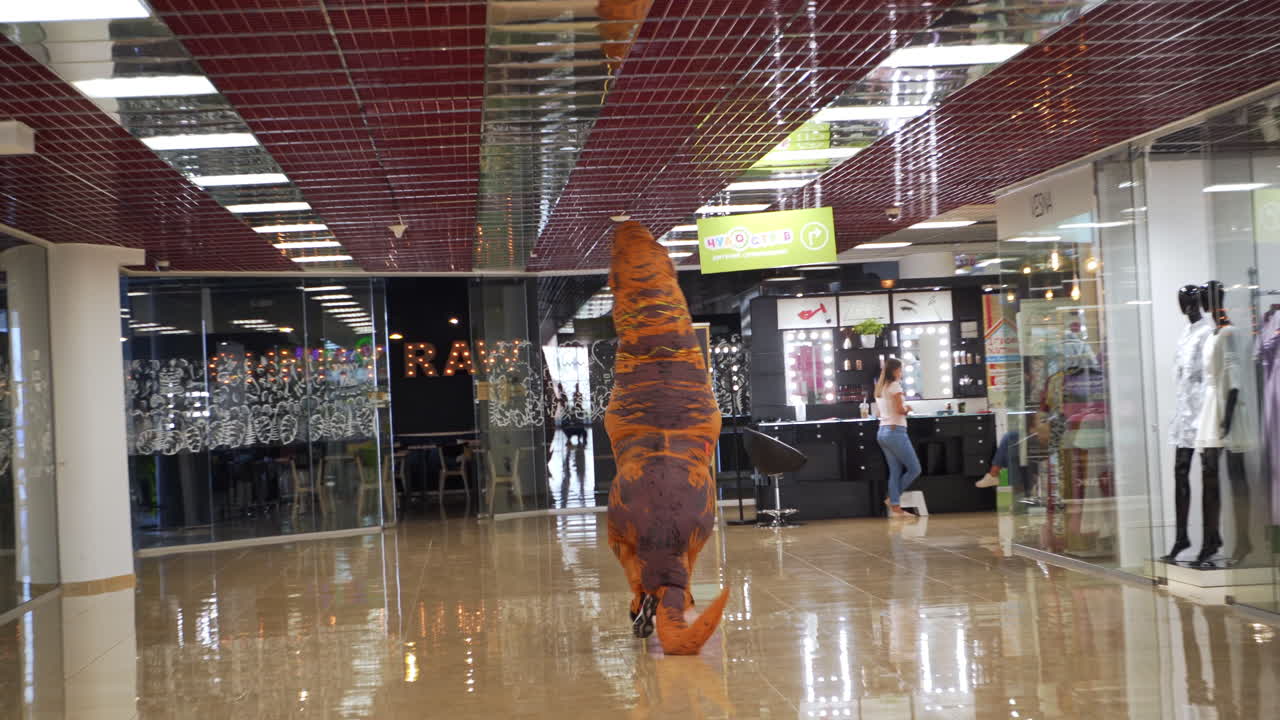 Man in orange funny spotty costume of dinosaur leaves the clothes shop. T-Rex runs quickly by the spacious hall of modern shopping mall.