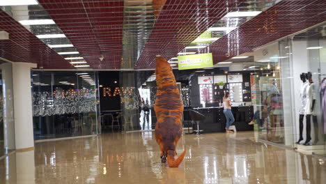 man in orange funny spotty costume of dinosaur leaves the clothes shop. t-rex runs quickly by the spacious hall of modern shopping mall.